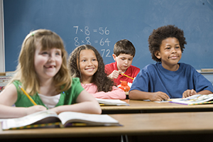 Children sitting at desks with books in classroom. Boy in background not paying attention.